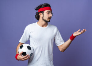 unpleased young sporty man wearing headband with wristband holding ball holding out hand at side isolated on blue background
